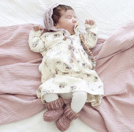 Baby girl in floral dress with knitted booties and bonnet, with dummy in mouth lying on dusty pink wrap