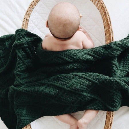 Baby lying on tummy in bassinette with forest knitted blanked draped over