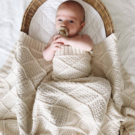Baby lying in bassinette with dummy in mouth and cream knitted blanket draped over